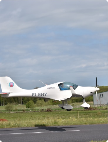 A small white aeroplane with the registration EI-EHY is taking off or landing on a runway surrounded by green grass and trees under a partly cloudy sky.