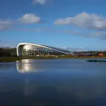 A modern, curved glass building with a white frame sits beside a calm lake, reflecting in the water. The sky is blue with scattered clouds, and trees line the background.