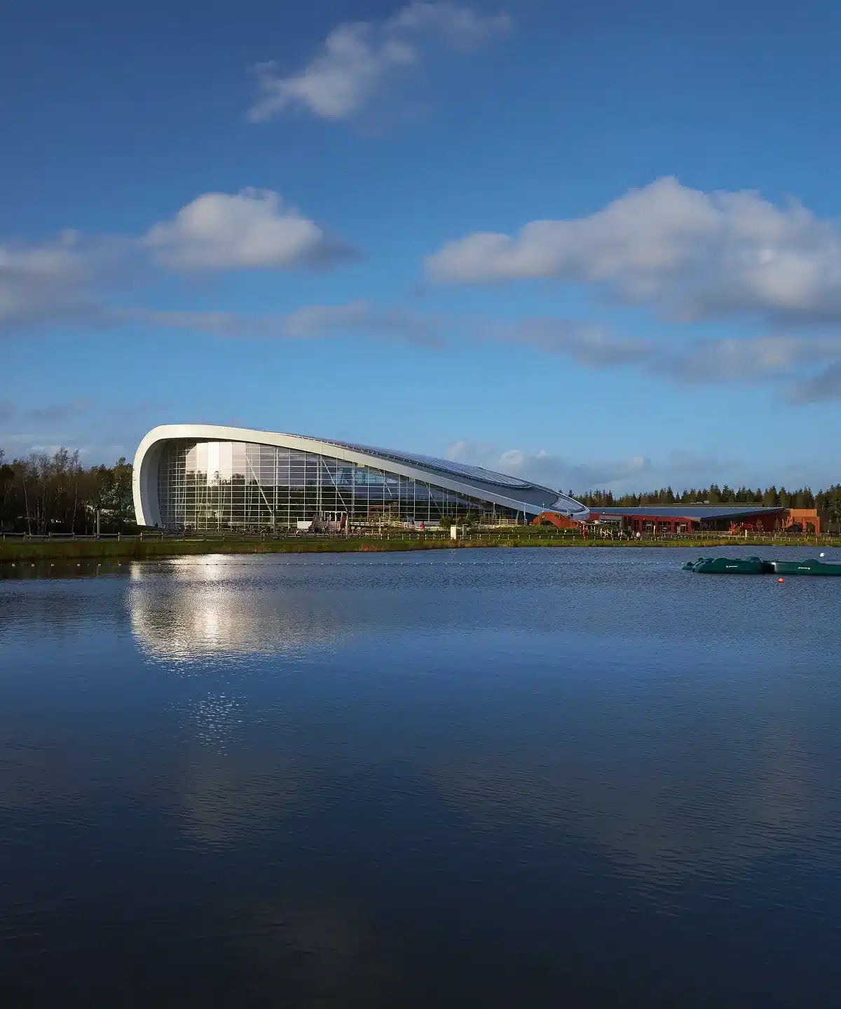 A modern, curved glass building with a white frame sits beside a calm lake, reflecting in the water. The sky is blue with scattered clouds, and trees line the background.