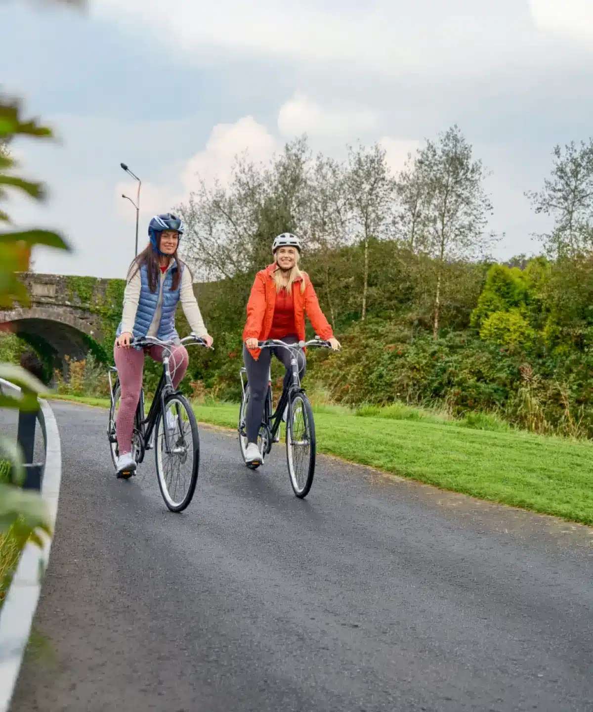 Two women riding bicycles on a paved path through a park, surrounded by green grass and trees, both wearing helmets and casual outdoor clothing.