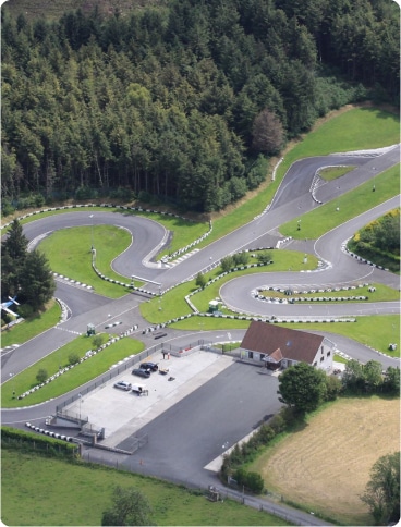 Aerial view of a winding go-kart or racing track surrounded by greenery and trees, with a car park, a small building, and parked vehicles near the entrance.