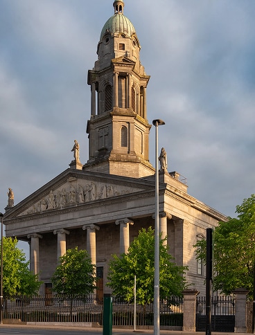 A neoclassical stone building with tall columns, statues on the roof, and a prominent central dome, surrounded by green trees and streetlamps under a partly cloudy sky.