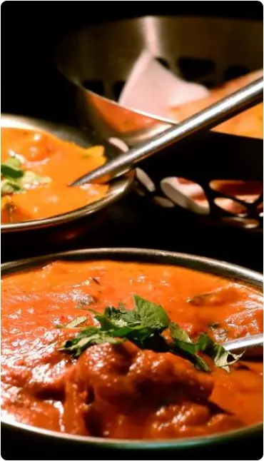 A close-up of two metal bowls filled with creamy orange Indian curry, garnished with fresh coriander. A serving spoon rests in the background, next to another dish. The food looks rich and flavourful.