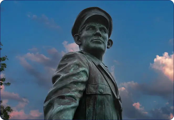 A bronze statue of a man in a military uniform and cap is seen from a low angle against a blue sky with clouds at sunset.