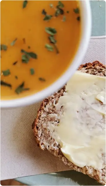 A bowl of orange soup garnished with herbs sits next to a slice of buttered wholegrain bread on a serviette.