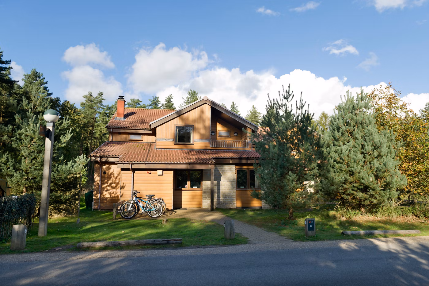 A two-storey wooden house with a red-tiled roof, surrounded by pine trees. Two bicycles are parked on the grass in front, and sunlight casts shadows on the building. A paved path leads to the entrance.
