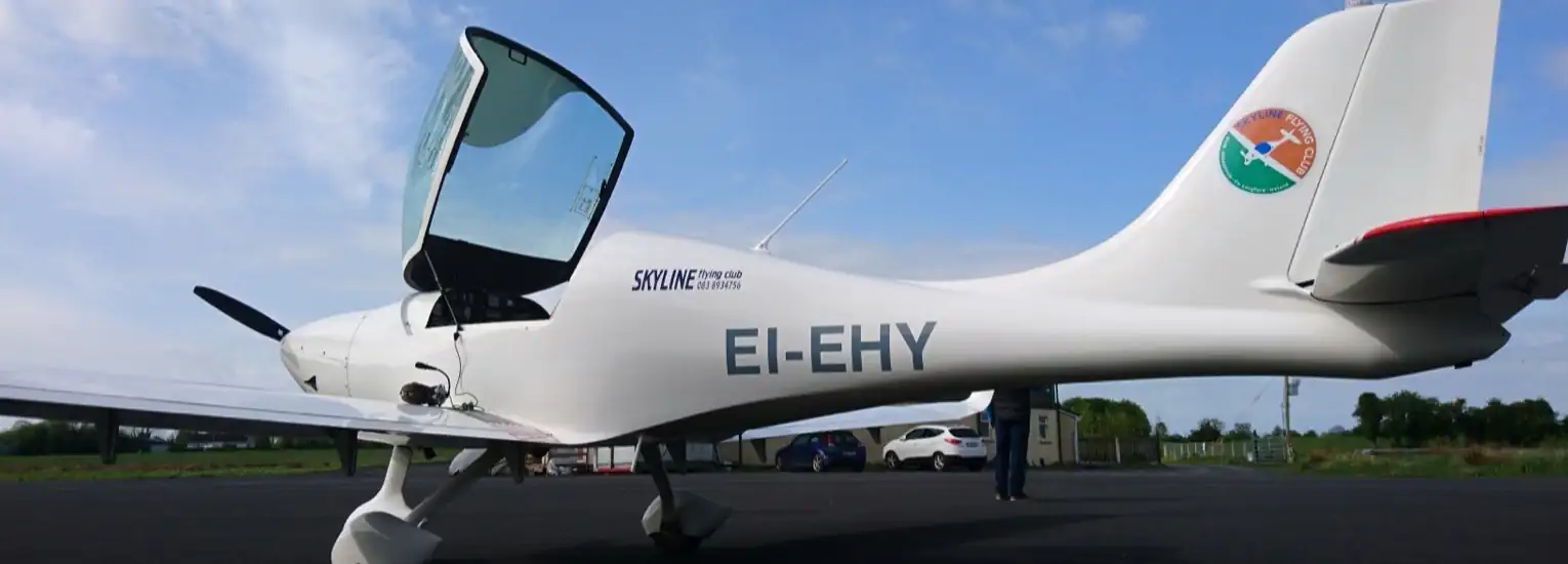 A small white light aeroplane with the registration EI-EHY is parked on a tarmac under a blue sky. The cockpit door is open and there are a few cars and greenery in the background.