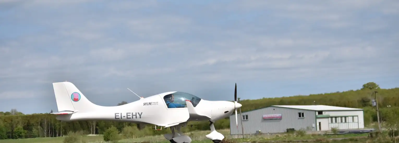 A small white single-engined aeroplane is taking off or landing on a runway, with a building and green trees in the background under a cloudy sky.