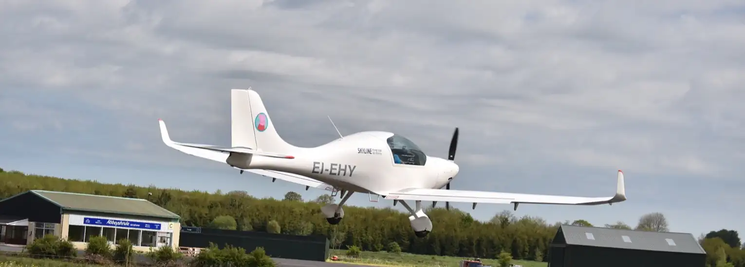 A small white single-engine aeroplane is taking off or landing on a runway at a rural airfield with green trees and buildings in the background under a cloudy sky.