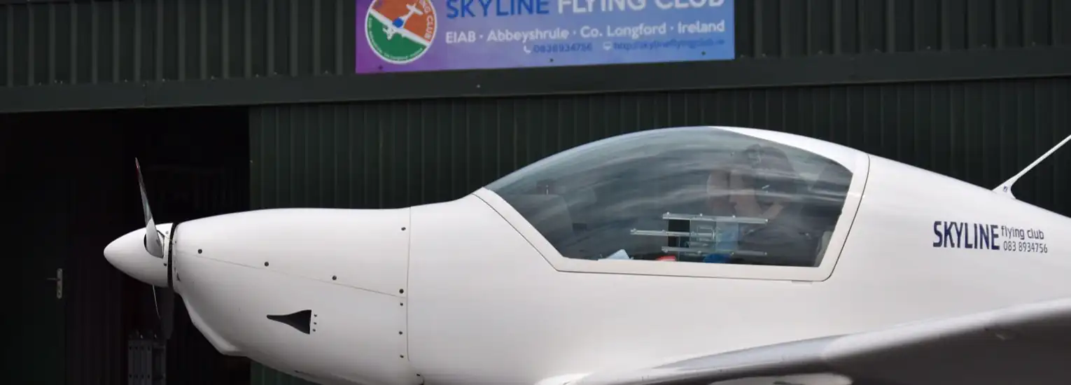A white single-engined aeroplane marked “SKYLINE flying club” is parked in front of a green hangar with a sign reading “SKYLINE FLYING CLUB” at Abbeyshrule, County Longford, Ireland.