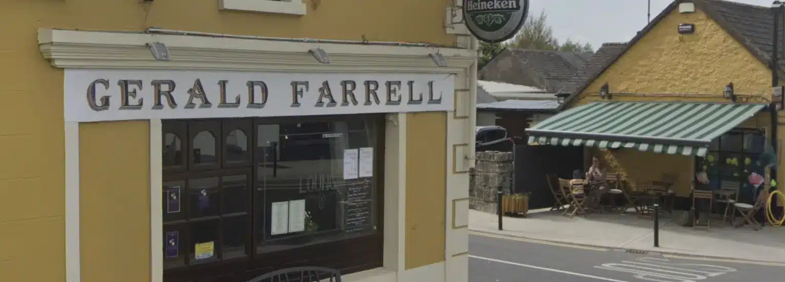 A yellow building with a sign reading "Gerald Farrell" above dark windows; across the street is a café with green-and-white striped awning and outdoor seating, with a woman sitting at a table.