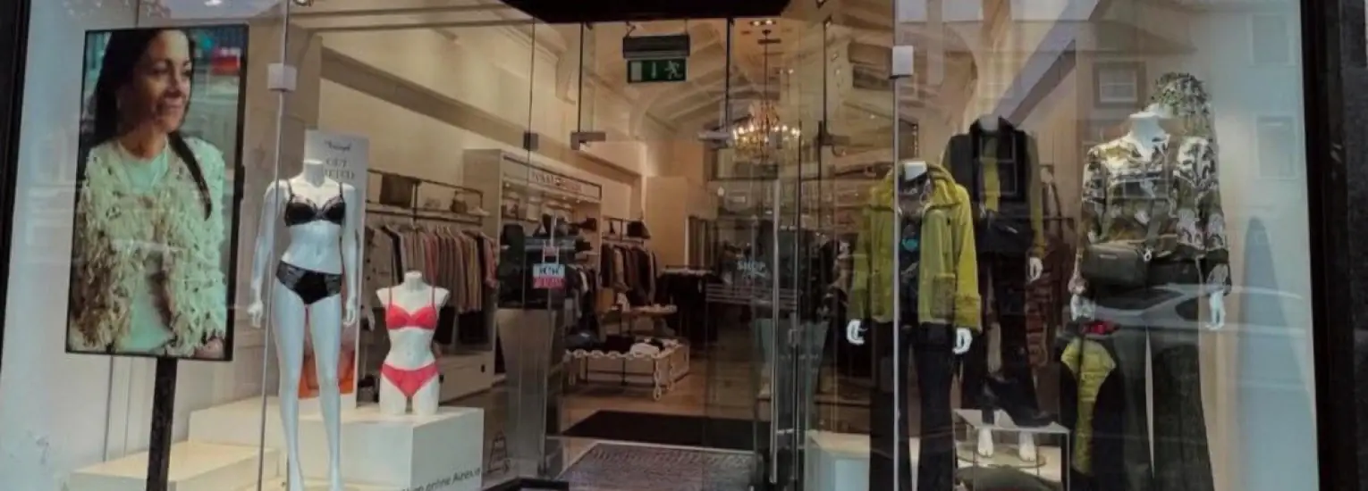 A clothing shop display with two mannequins in lingerie, two mannequins in casual outfits, and a digital screen showing a woman’s portrait. Glass doors reveal more clothing and a chandelier inside.
