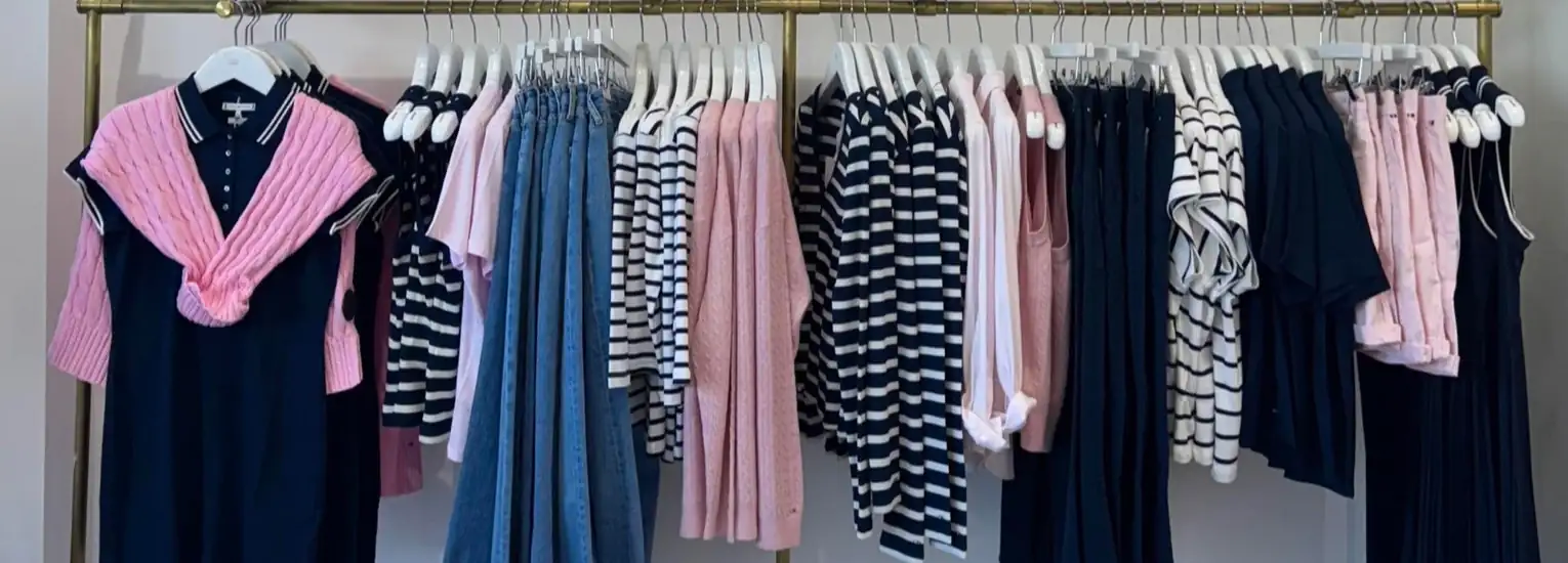 A clothes rail displays neatly hung garments, including striped and plain navy, pink, and blue tops and dresses, as well as blue jeans. A pink jumper is draped over a navy dress on the left.