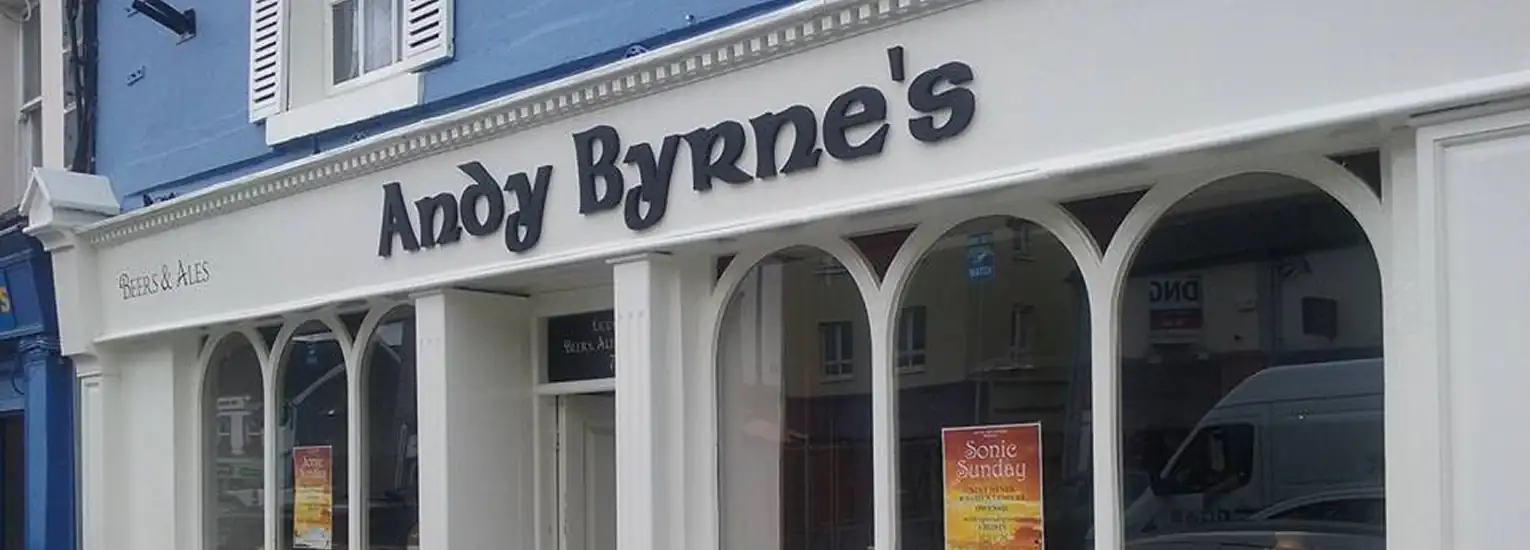 Shopfront with a white exterior and large arched windows. The sign above the windows reads "Andy Byrne's" in bold, black letters. Posters advertising events are visible in the windows.