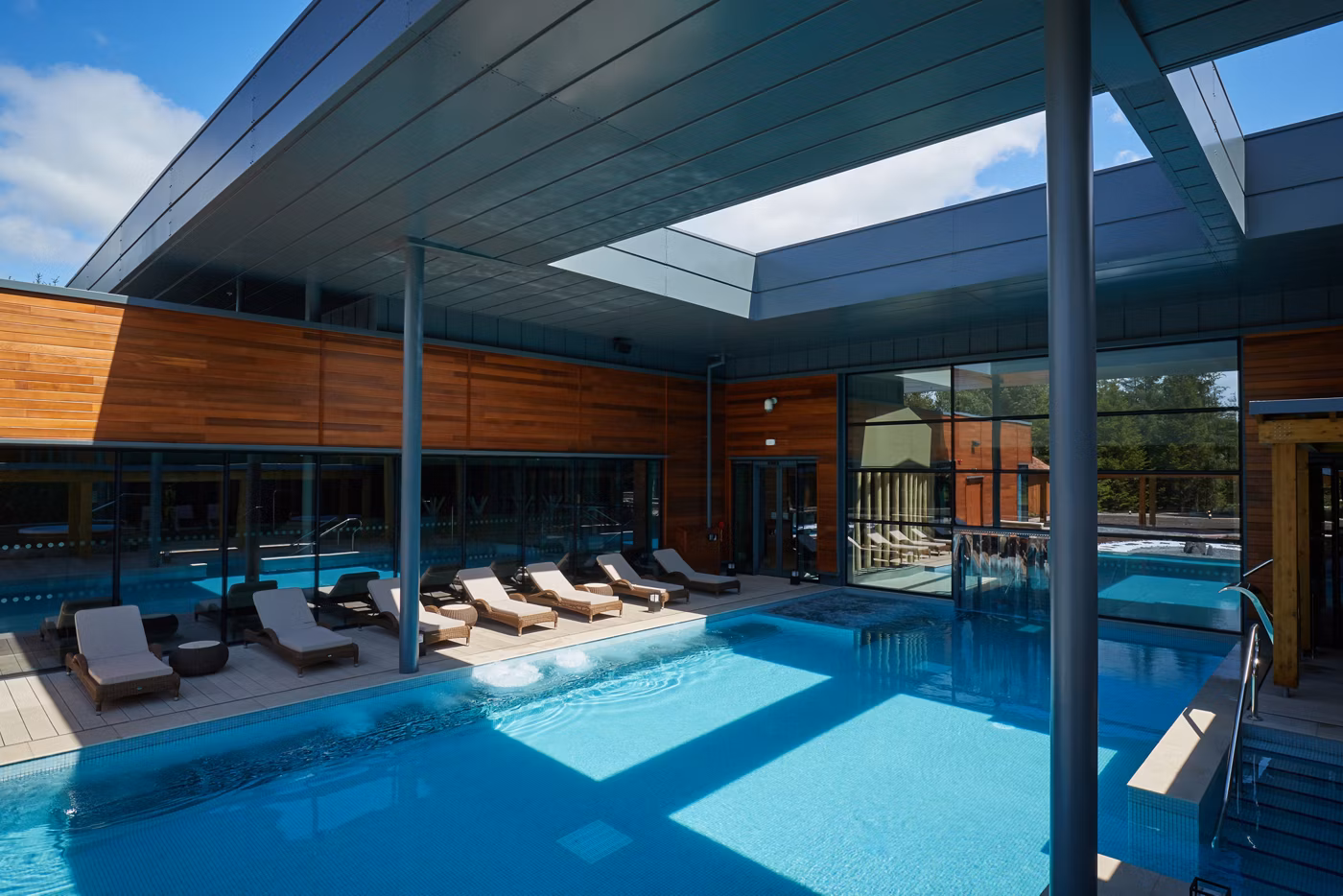 Modern outdoor pool area with several loungers beside a clear blue swimming pool, surrounded by a building with large windows and timber-panelled walls, under a partly covered roof.
