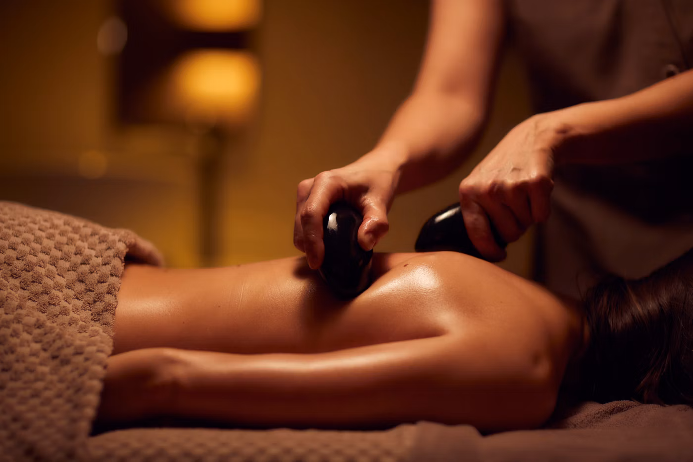 A person receives a hot stone massage, lying face down on a massage table covered with a textured towel in a softly lit, relaxing spa environment.