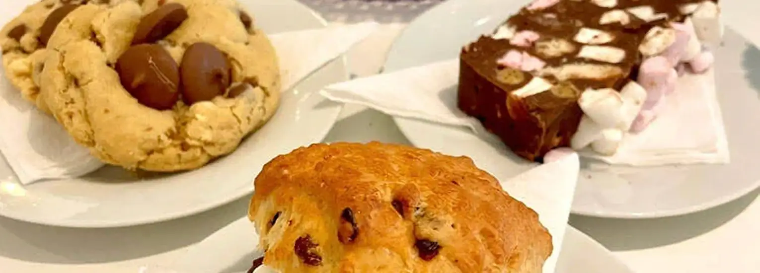 Three dessert plates on a table: a large biscuit with chocolate pieces, a chocolate bar with marshmallows, and a golden sultana scone with visible raisins, each served on a white serviette.