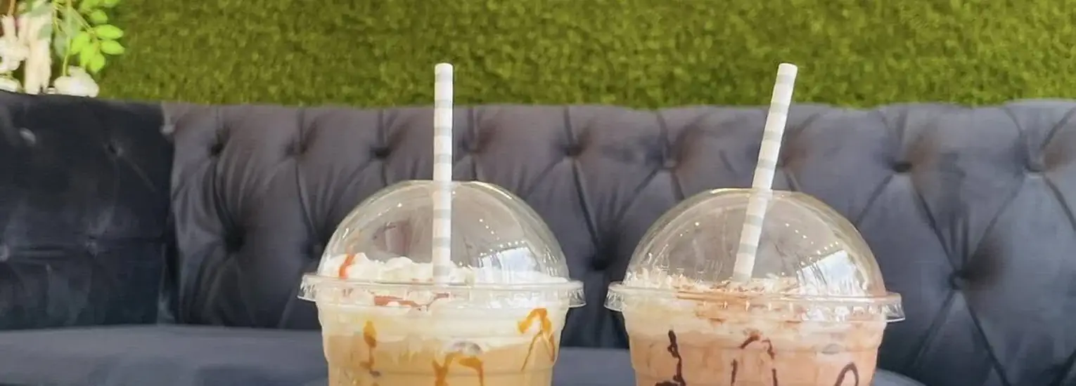 Two iced drinks with whipped cream, caramel, and chocolate drizzle sit on a dark blue tufted sofa, with a green artificial grass wall in the background. Both drinks have domed lids and striped straws.