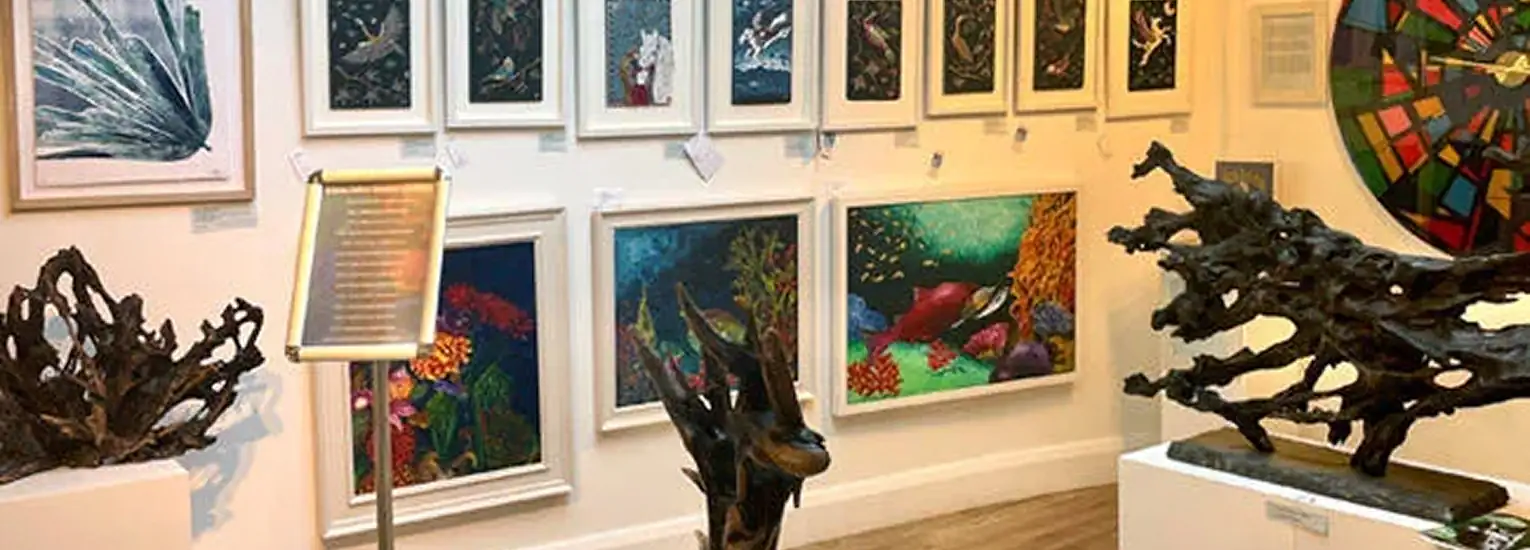Art gallery with colourful paintings of underwater scenes and coral reefs on the walls, and dark, abstract sculptures displayed on white plinths in the foreground.