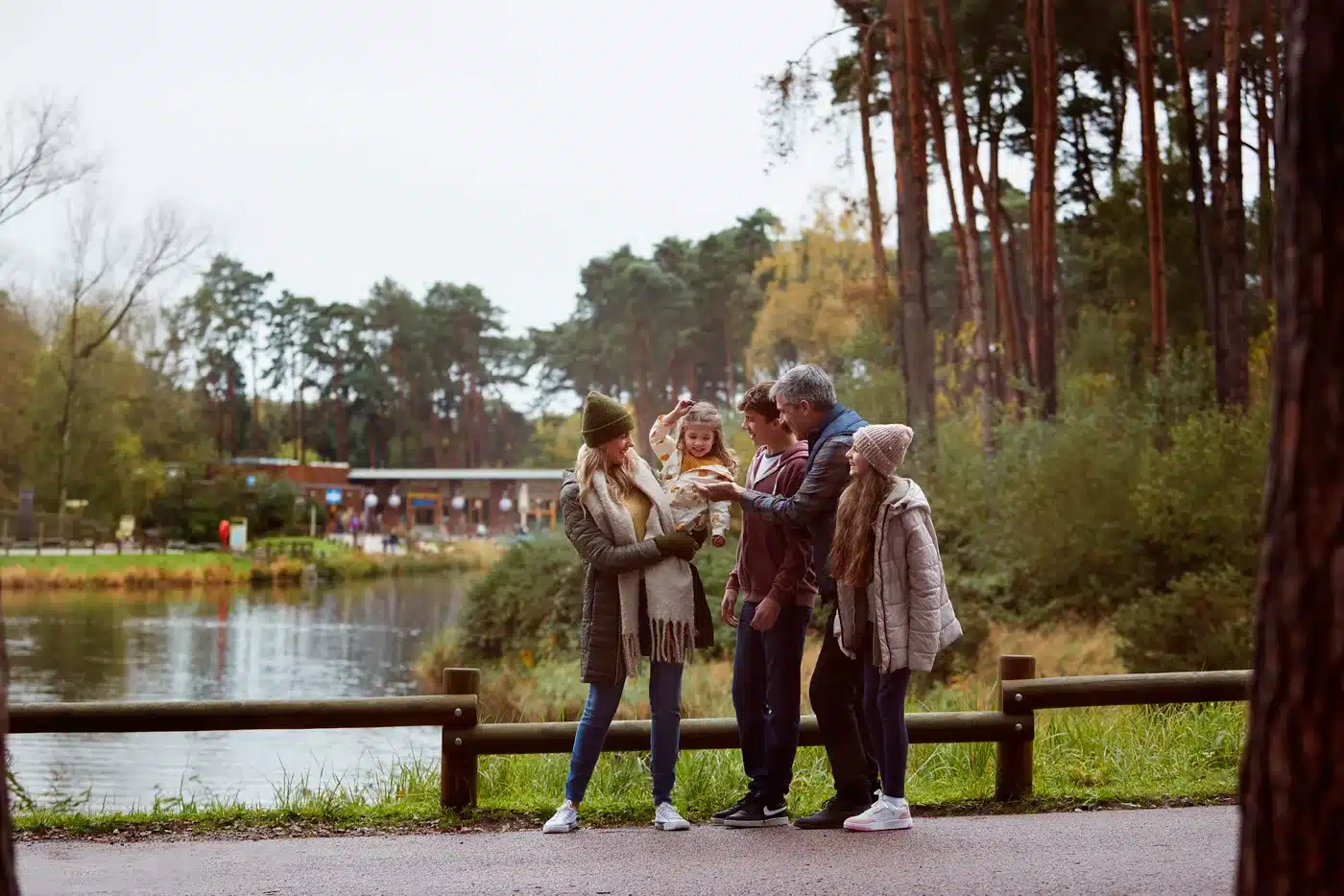 A family of four, dressed warmly in coats and hats, stands together by a lake in a wooded park. The parents are holding a young child whilst another child stands beside them. Trees and water are in the background.