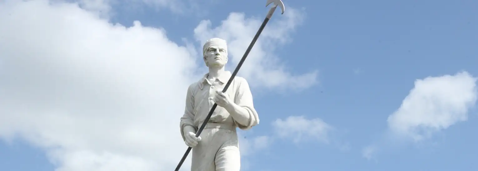 A white statue of a person holding a long pole with a hook stands against a partly cloudy blue sky.