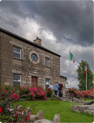A stone building with a clock above the door and two flags outside, one Irish and one French. Three people stand talking near the entrance, and pink flowers bloom in the garden. The sky is cloudy and overcast.