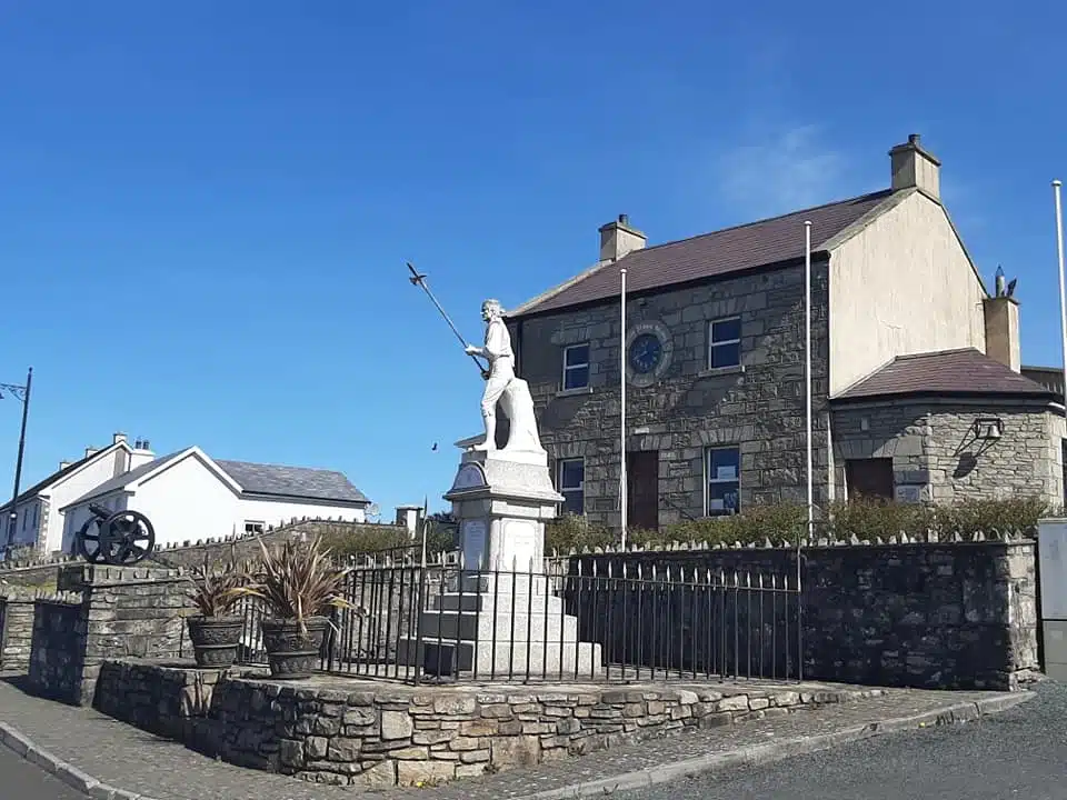 A stone statue of a figure holding a trident stands on a plinth in a small fenced area. Behind it is a two-storey stone building with a clock on its façade, under a clear blue sky.