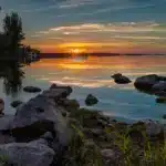 A calm lake at sunset—with rocks in the foreground and trees on the left—offers a perfect Outdoor Adventure Longford moment, as the glowing orange sun nears the horizon and clouds reflect in the still water.