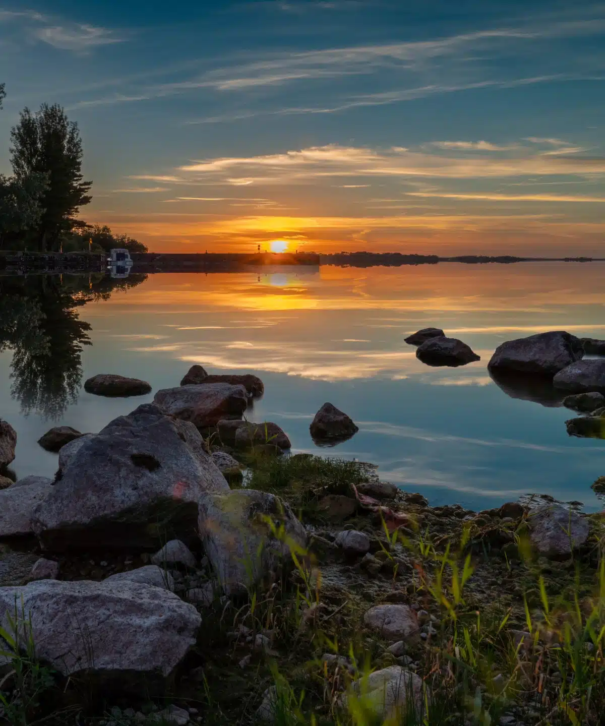 A calm lake at sunset—with rocks in the foreground and trees on the left—offers a perfect Outdoor Adventure Longford moment, as the glowing orange sun nears the horizon and clouds reflect in the still water.