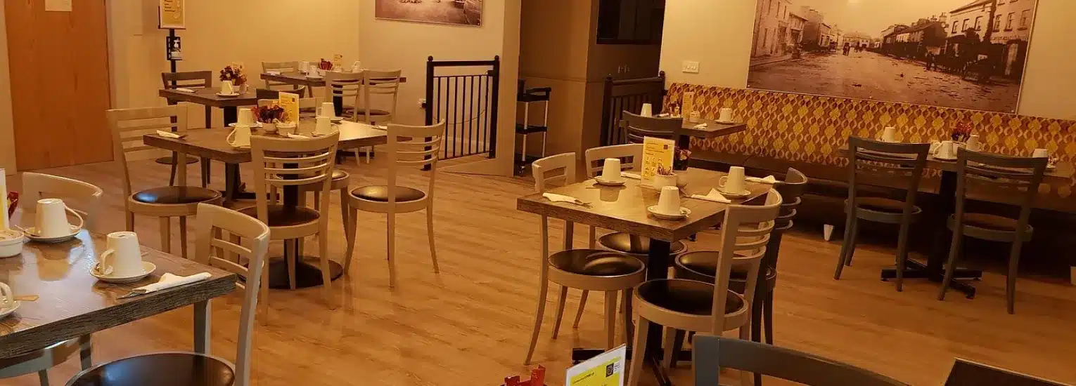 A cosy, well-lit café with wooden floors, several empty tables and chairs, cups and menus on tables, and vintage black-and-white photos decorating the walls.