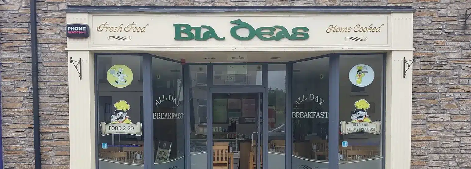Front view of Bia Deas café with large glass windows, stone exterior, and signs reading “Fresh Food,” “Home Cooked,” “All Day Breakfast,” and “Food 2 Go.” Cartoon chef images are displayed on the windows.