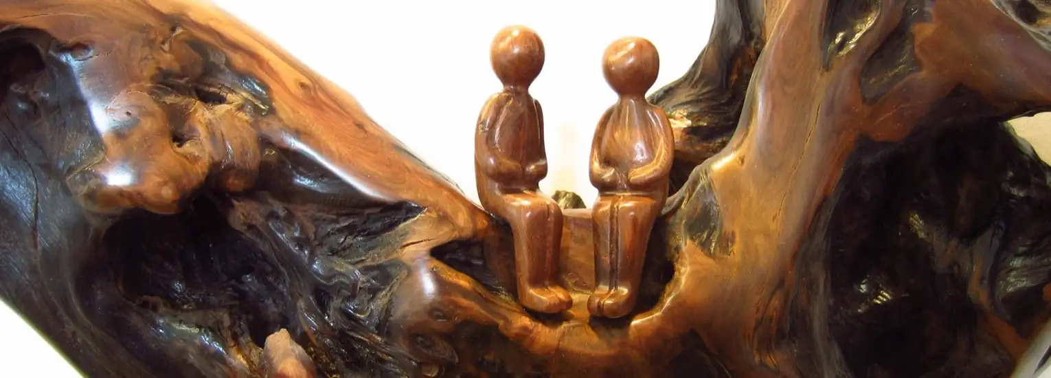 Two small, simple wooden figurines sit side by side on a polished, abstract piece of wood, resembling a natural bench or branch. The figures appear to be engaged in quiet companionship.