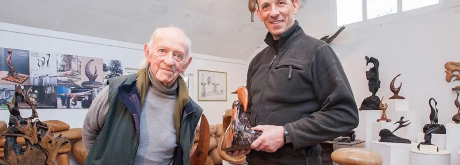 Two men stand in an art studio surrounded by abstract wooden sculptures. One man holds a small sculpture, and both are smiling at the camera. The walls display more artworks and photos. Natural light enters from a window.