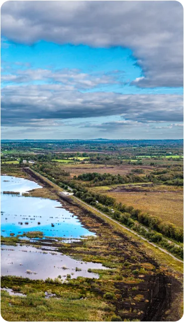A landscape view of a peat bog with patches of water and muddy tracks under a cloudy sky, surrounded by fields and greenery in the distance.