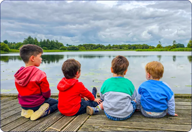 Four young boys sit side by side on a wooden jetty, facing a calm lake surrounded by greenery under a cloudy sky. They wear colourful hoodies and appear to be enjoying the peaceful scenery.
