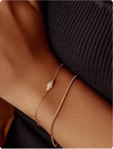 A close-up of a person's wrist wearing two delicate bracelets, one with a small diamond-shaped charm and the other a simple gold band, against a dark ribbed fabric background.