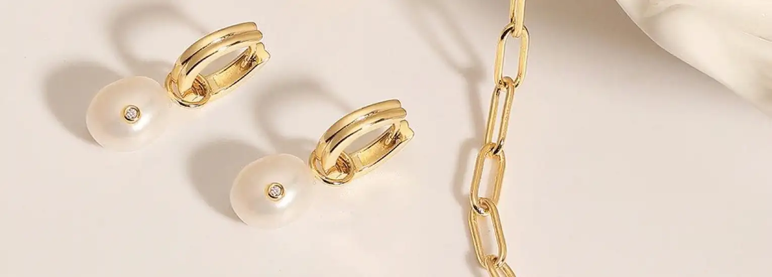 Gold hoop earrings with white pearl and diamond accents are placed beside a gold chain link necklace on a light beige surface. A soft white fabric is visible in the top right corner.