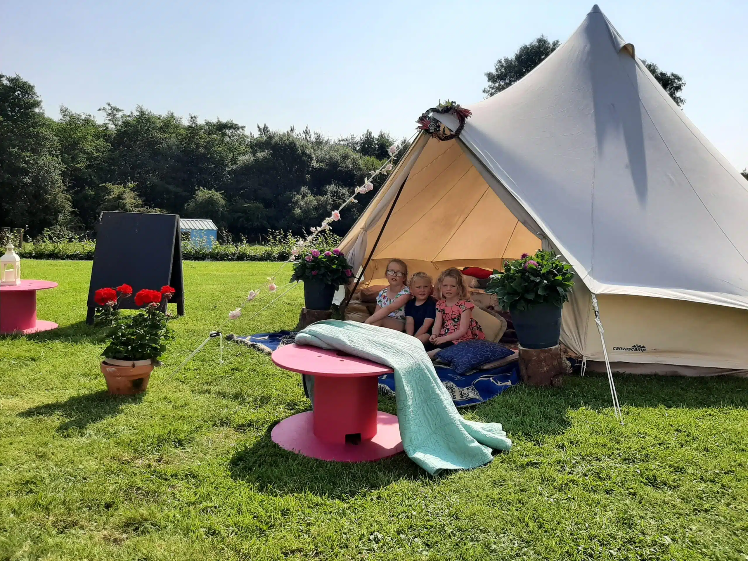 Three children sit inside a cream-coloured bell tent on grass, surrounded by potted flowers and pink tables. A cosy blanket is spread in front, with trees and a chalkboard in the background under a sunny sky.