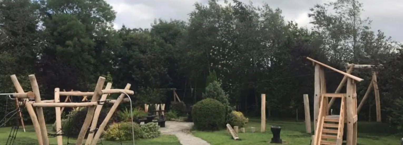 Wooden playground equipment, including climbing frames and a zip wire, is set on a grassy area with a winding path. Trees and shrubs surround the play area under a cloudy sky.