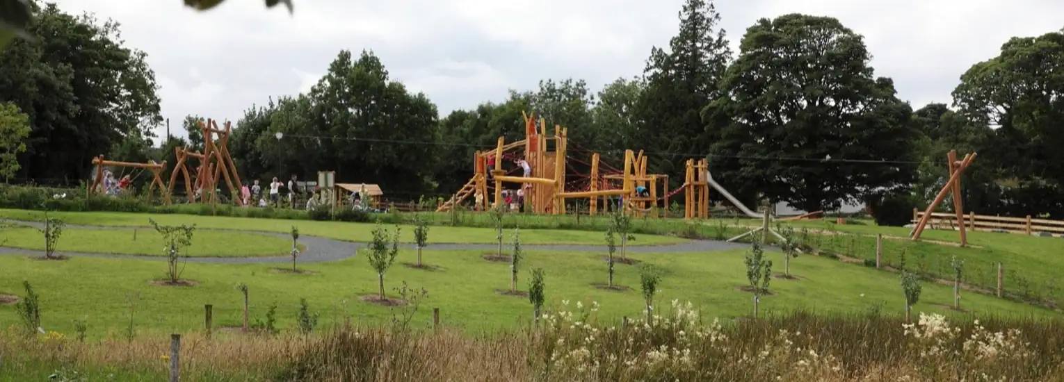 A large wooden playground structure surrounded by grass, trees, and walking paths, with children and adults playing and walking in the park on a cloudy day.