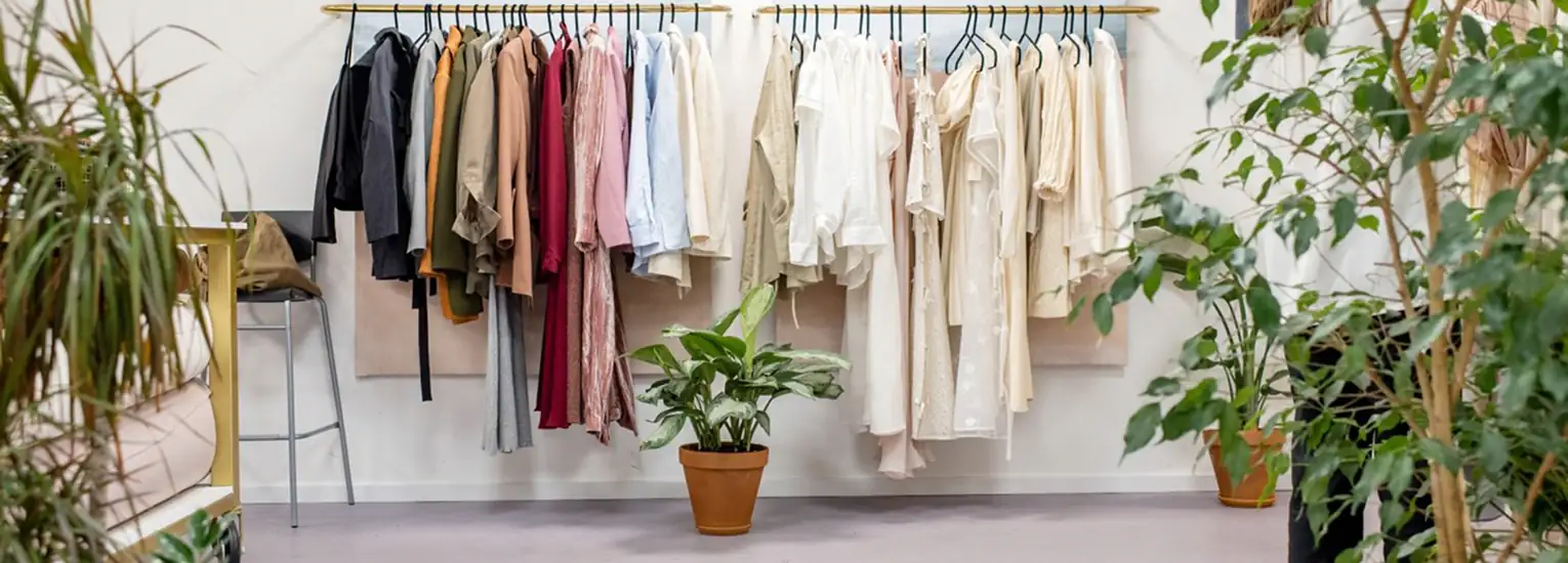 A clothes rail displays various shirts, blouses, and jackets in neutral tones, flanked by green potted plants against a white wall in a minimalist room.