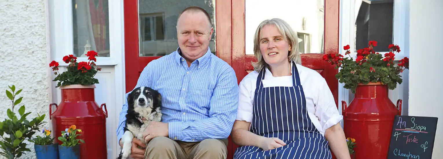 A man and woman sit on a doorstep surrounded by red milk churns and potted geraniums. The man, in a blue shirt, holds a black and white dog. The woman wears a chef’s jacket and striped apron. A chalkboard sign is on the right.