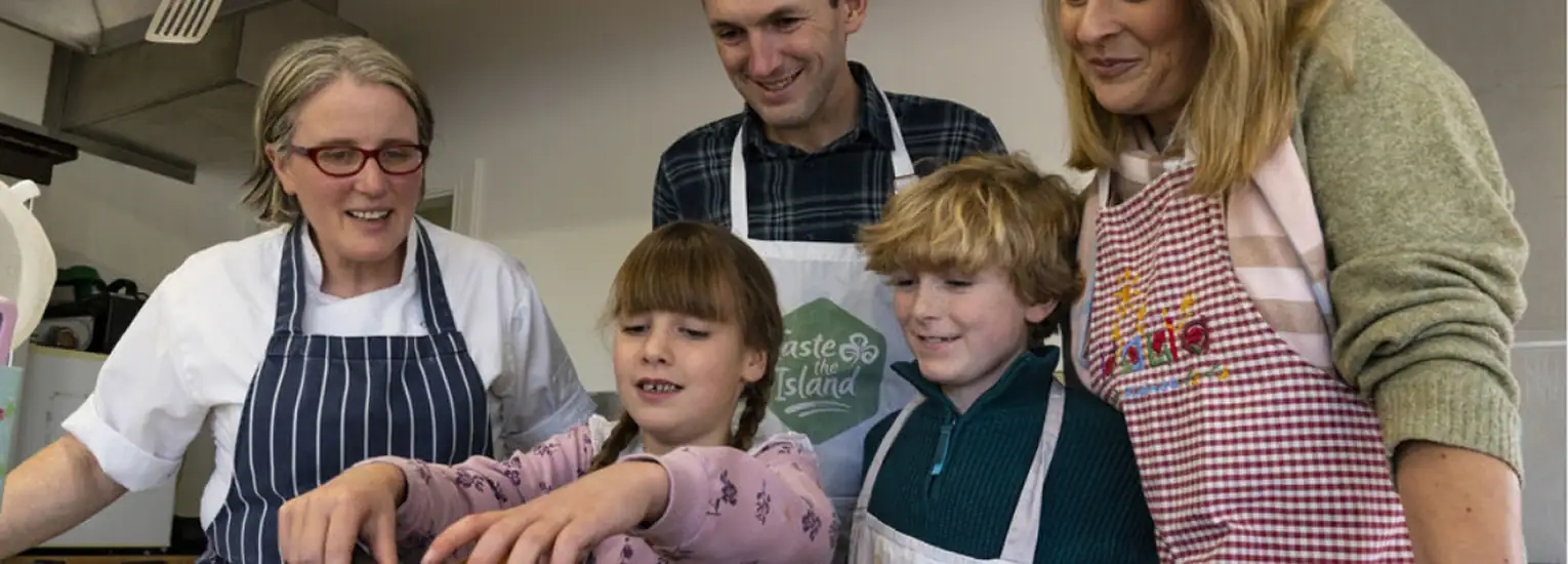 A group of five people, including two children and three adults wearing aprons, gather around a kitchen worktop, smiling and preparing food together in a bright kitchen.