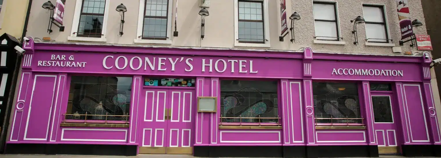 The front of Cooney's Hotel, painted bright pink with white trim, features signage for a bar, restaurant, and accommodation. Large windows and several lamps are visible on the building’s façade.