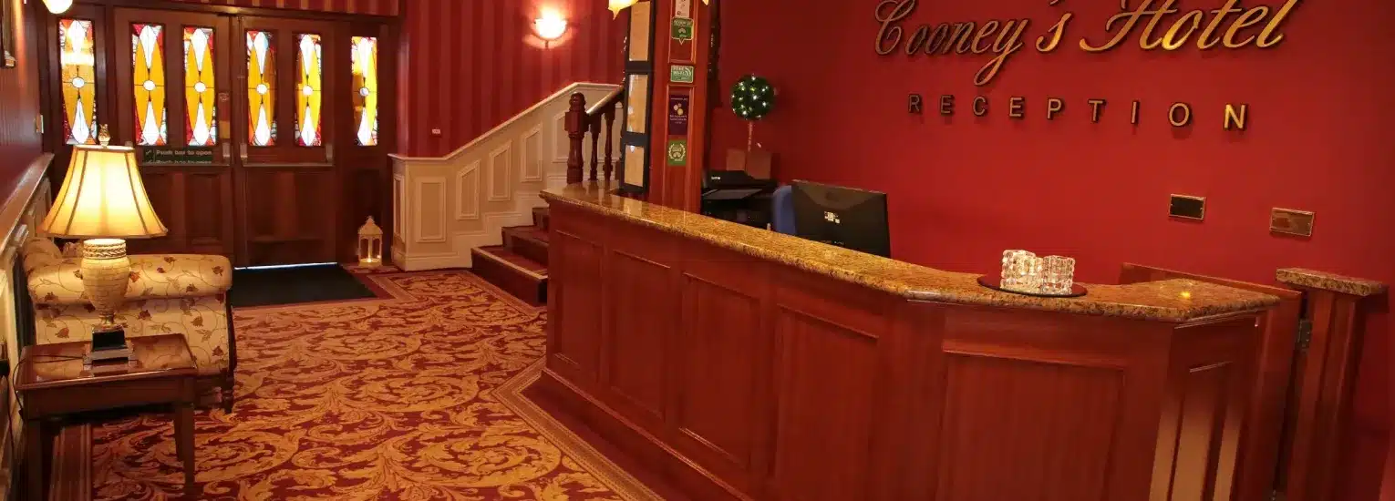 A warmly lit hotel reception area with a wood-panelled front desk, patterned carpet, floral sofa, and stained glass entry doors; the wall reads “Convey’s Hotel Reception” in gold letters.