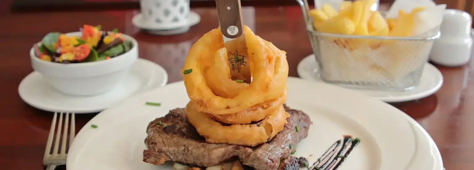 A grilled steak topped with onion rings and a knife, served on a white plate. A bowl of salad and a basket of chips are on the side, all set on a wooden table.