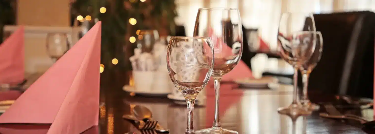 A close-up of a restaurant table set with folded pink serviettes, clear wine glasses, cutlery, and plates. Warm lighting and a blurred background with fairy lights create an inviting, elegant atmosphere.