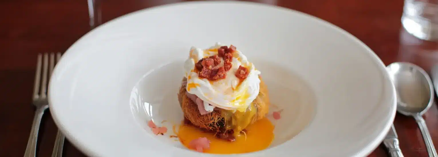 A poached egg with a runny yolk sits on toasted bread, topped with small pieces of bacon, served on a white plate with a fork and spoon on either side.