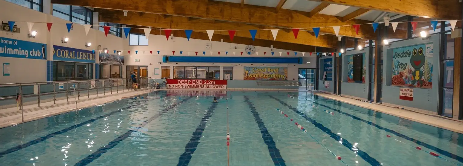 Indoor swimming pool with lane dividers, safety signs, and colourful bunting hanging above. Walls display murals and posters. Large windows and ceiling beams are visible, and pool rules are posted at the deep end. The area appears empty.