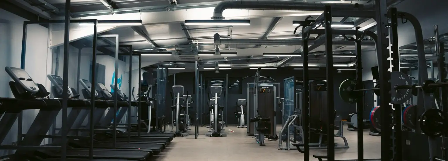 A modern gym interior with treadmills lined up on the left, various weight machines on the right, and strength training equipment in the centre under bright overhead lights.